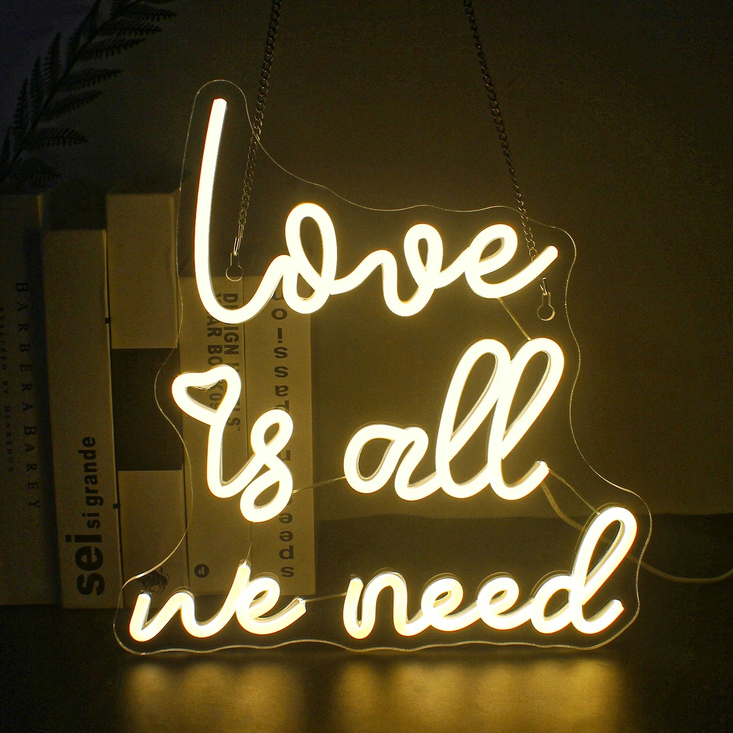 A imagem apresenta uma vibrante placa de LED neon que diz "O amor é tudo o que precisamos" em uma fonte fluida e cursiva.