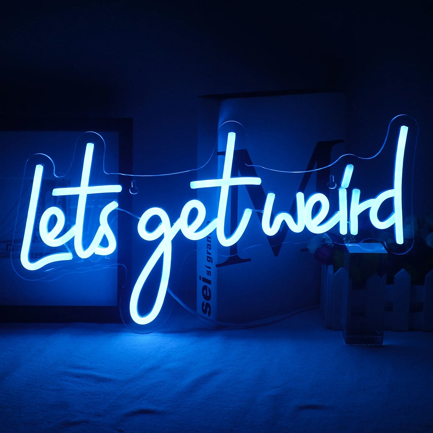 A imagem mostra uma vibrante placa de LED neon com os dizeres "Let's Get Weird" (Vamos Ficar Estranhos) montada em uma parede.