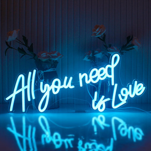 A imagem exibe uma vibrante placa de LED neon que diz a frase "All You Need Is Love" em uma fonte cursiva e estilosa.