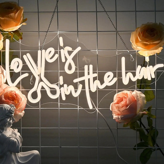 The image features a vibrant, handmade LED neon sign spelling out the phrase "Love is in the Hair.