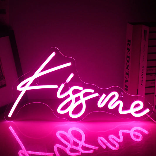 A vibrant "Kiss Me" LED neon sign is illuminated, displaying bright and bold pink lettering against a dark background.