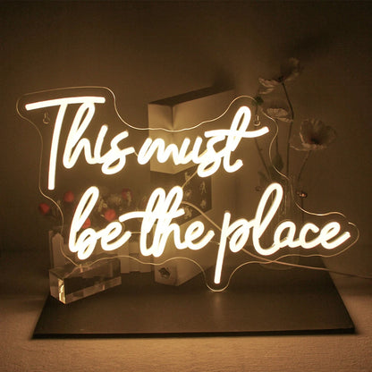 The image displays a vibrant LED neon sign that reads "This must be the place" in cursive script.