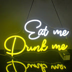 A imagem apresenta uma vibrante placa de LED neon exibindo as palavras "Eat Me Drink Me" em uma fonte cursiva e excêntrica.
