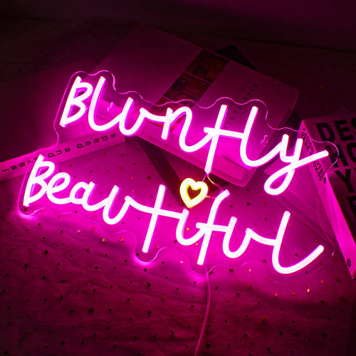 A imagem apresenta uma vibrante placa de LED neon feita à mão exibindo a frase "Bluntly Beautiful".