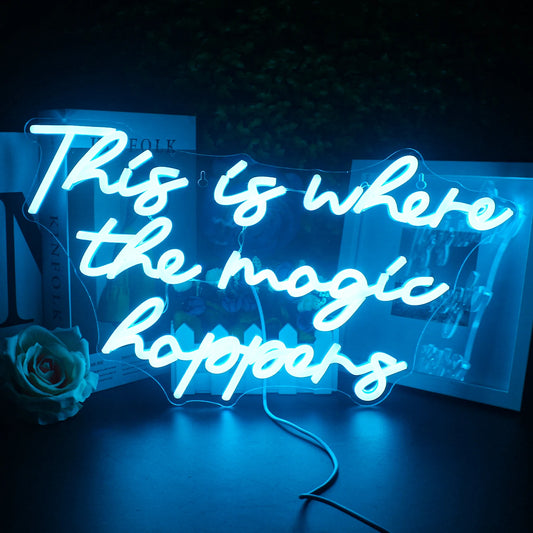 The image shows a vibrant neon sign illuminating the phrase "This is Where The Magic Happens" in a stylish, cursive font.