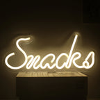 A imagem mostra uma vibrante placa de LED neon no formato da palavra "Snacks" em uma fonte cursiva e divertida.