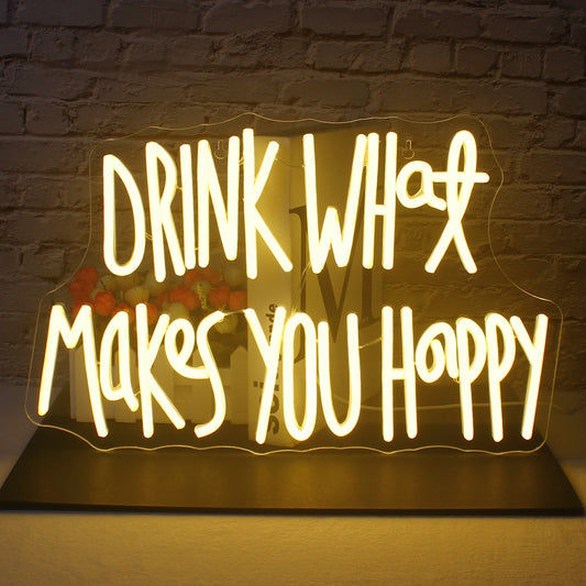 In the image, there is a vibrant LED neon sign displaying the phrase "Drink What Makes You Happy" in an elegant cursive font.