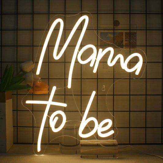 The image features a vibrant and stylish LED neon sign displaying the phrase "Mama to Be" in elegant, cursive lettering.