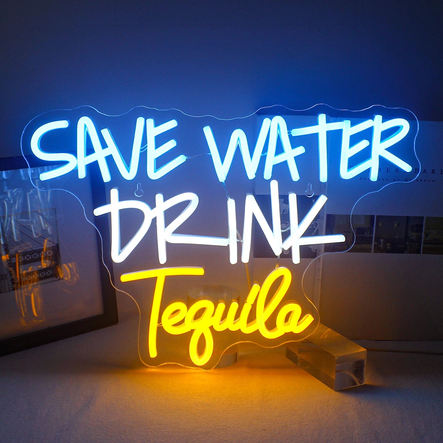 The image features a vibrant LED neon sign displaying the phrase "Save Water Drink Tequila" in bold, playful typography.