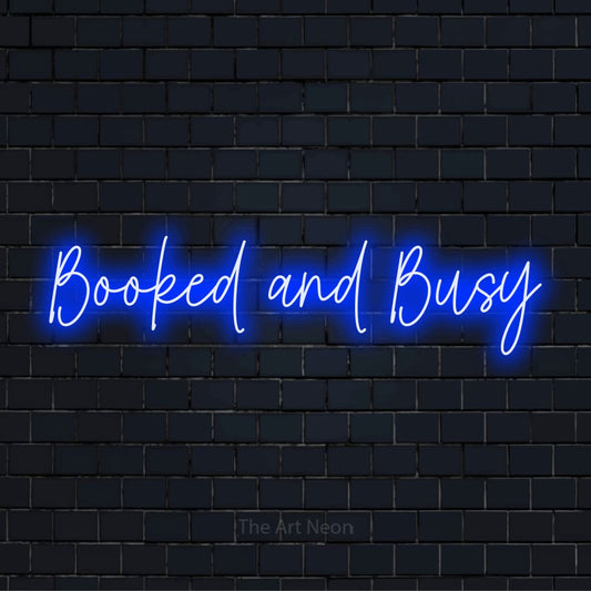 "Booked And Busy" glowing LED neon sign on a black brick wall