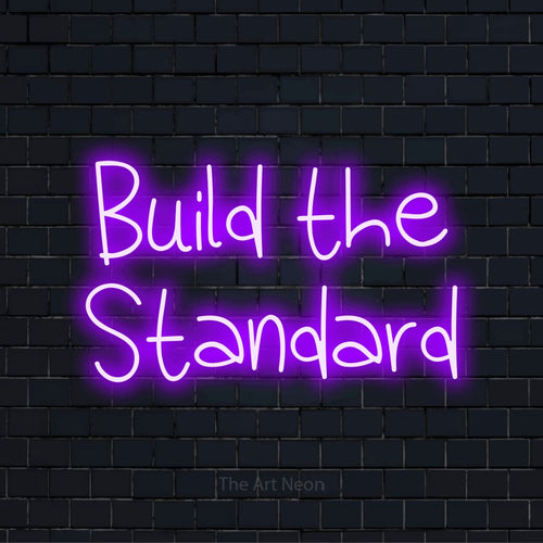 "Build The Standard" script-style LED neon sign on a black brick wall
