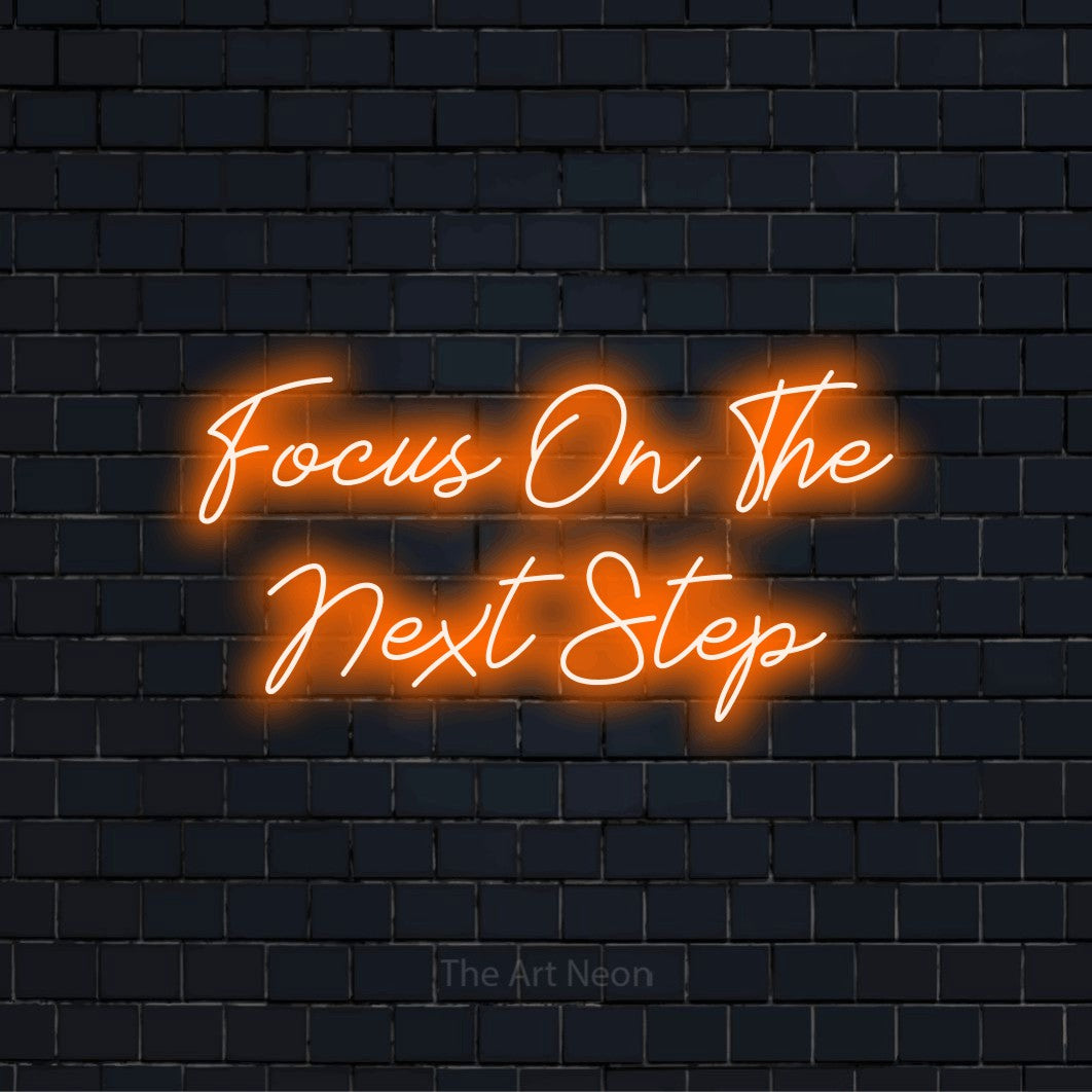 "Focus On The Next Step" script-style LED neon sign on a black brick wall