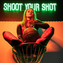 A person sitting inside a basketball hoop, holding a basketball, with a green neon LED sign that reads "Shoot Your Shot" glowing above them. The setup features a basketball-themed concept with vibrant green and orange lighting, creating an energetic and motivational atmosphere. This image highlights The Art Neon's custom neon signs for sports and inspiration themes