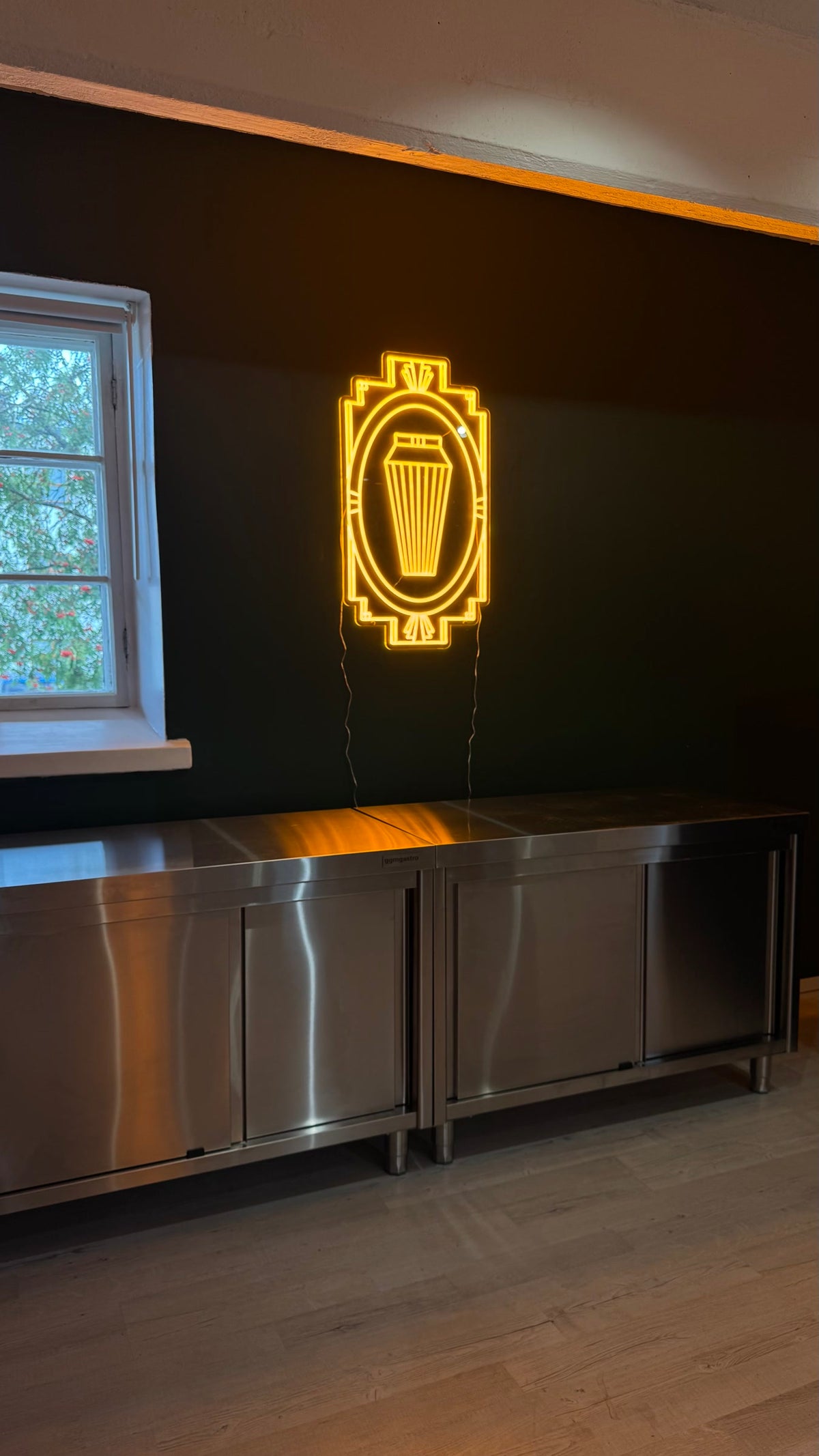 Ícone de shaker de cocktails em néon LED amarelo quente, numa moldura Art Déco sobre uma parede escura.