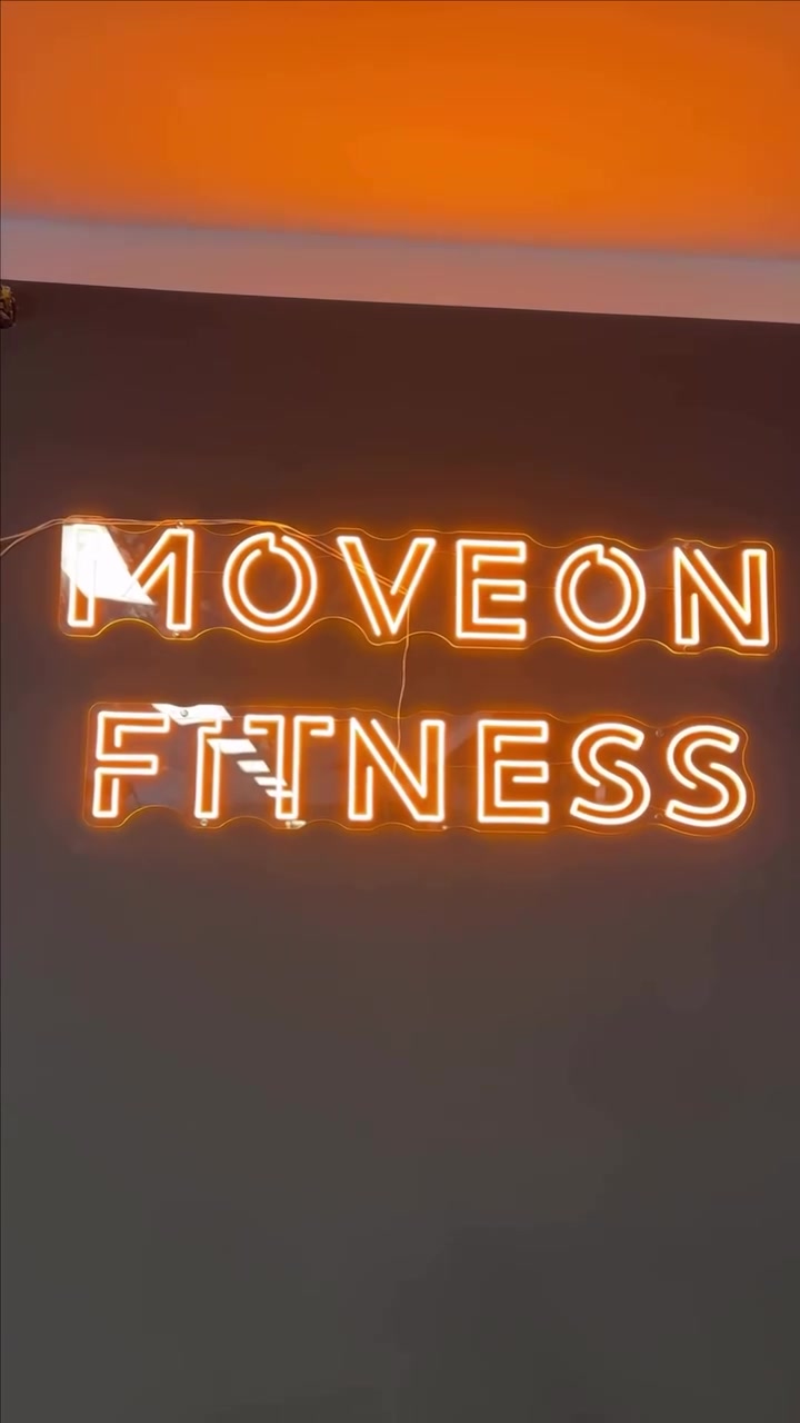 Warm-yellow MOVEON FITNESS LED neon logo mounted on a dark gym studio wall.